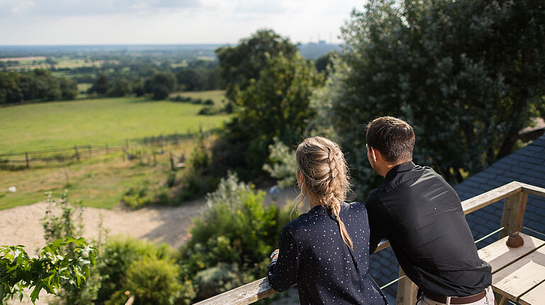un couple est accoudé sur un terrasse bois d'une maison qui surplombe des champs de pâturage avec haie bocagère. 